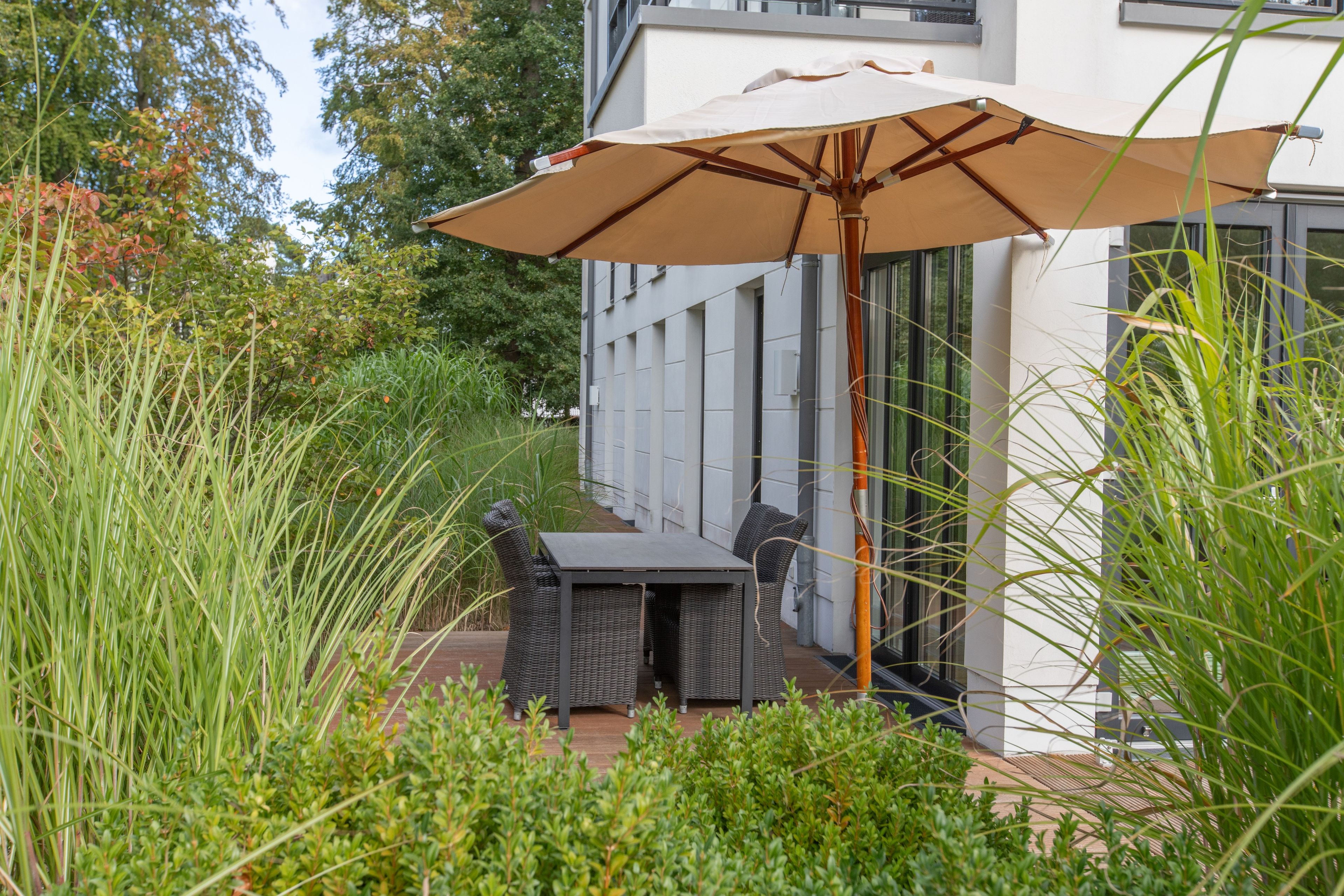 A patio with a table and chairs, surrounded by tall grass and a building in the background.