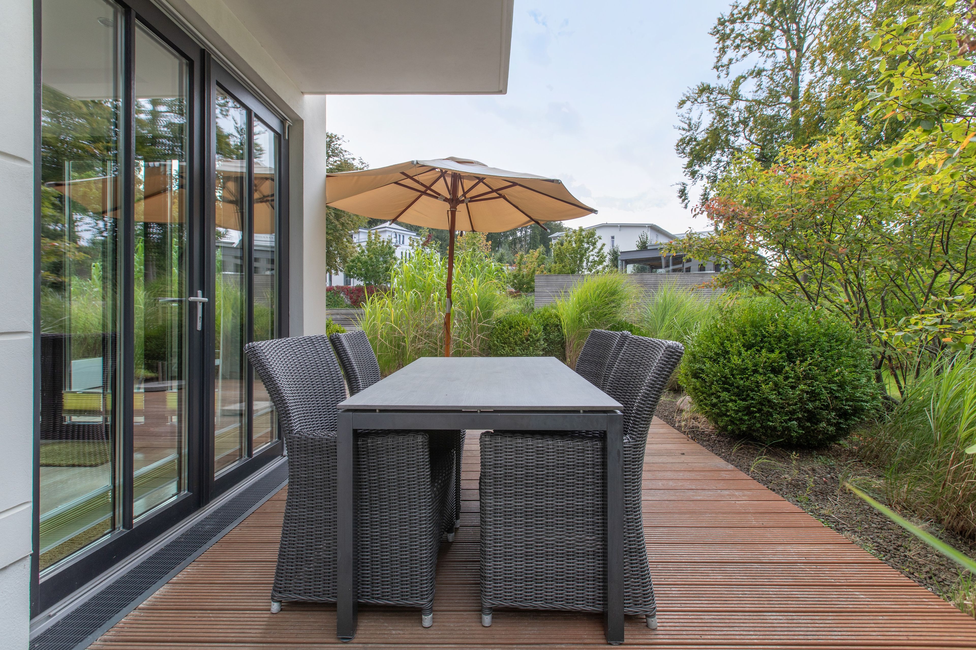 En udendørs patio med et bord, stole og en parasol, samt udsigt til grøn vegetation og bygninger.