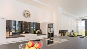 Une cuisine moderne avec un îlot central, une grande horloge murale et un panier de pommes posé sur le plan de travail.