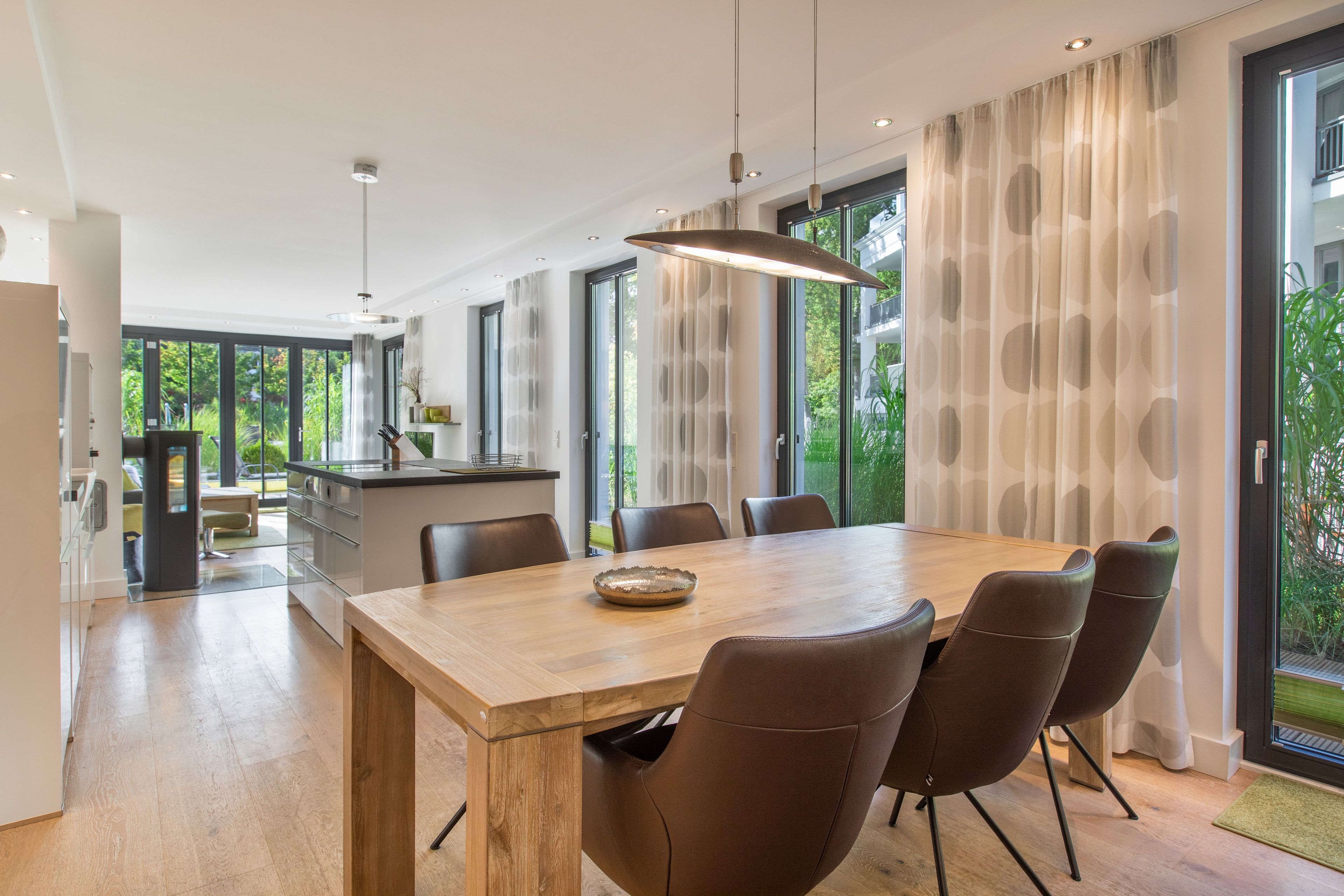 A modern dining area with a wooden table and leather chairs, large windows with curtains, and a kitchen in the background.