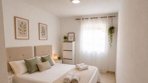 Ein Schlafzimmer mit Bett, Kissen, Nachttisch, Lampe, einem gerahmten Bild, einer Pflanze und einem Fenster mit Vorhängen.