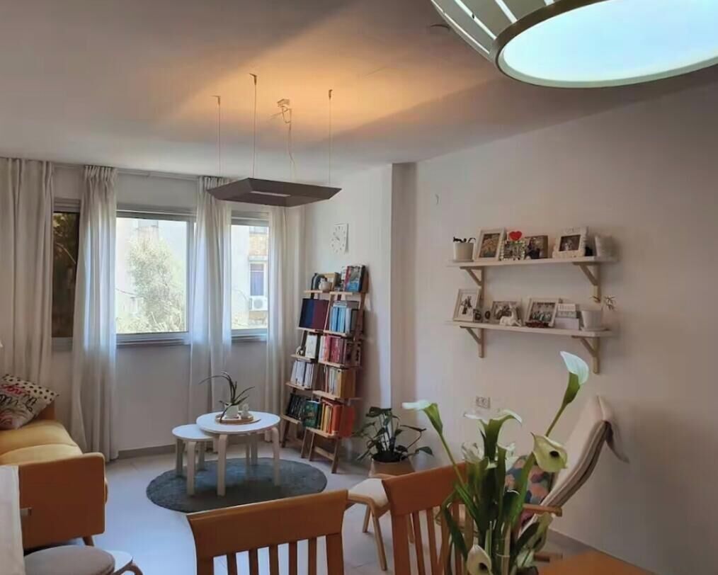 A living room with a sofa, a round table, a bookshelf, and a wall-mounted shelf with pictures.