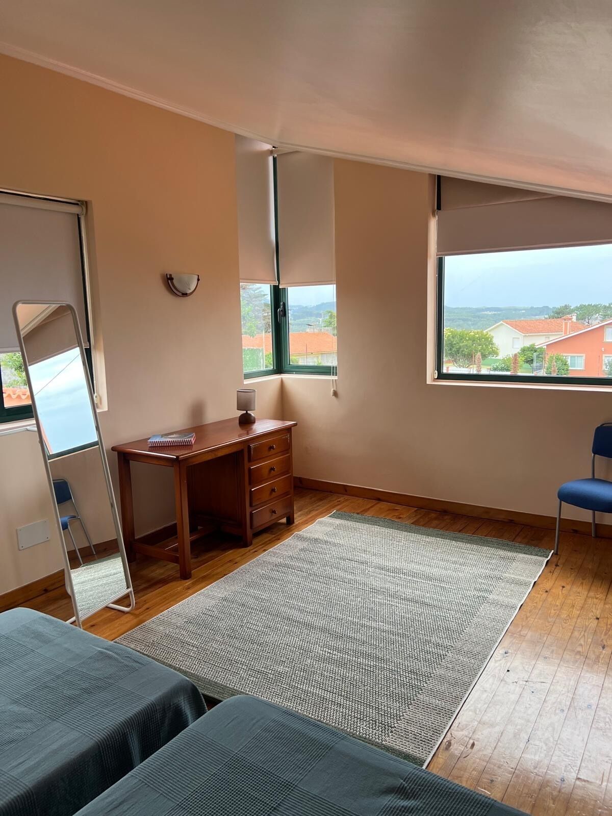 Ein Schlafzimmer mit einem Holztisch, einem Sessel, einem Stehspiegel und einem großen Fenster mit Blick auf Gebäude.