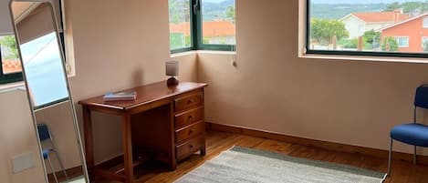 Un dormitorio con escritorio de madera, una silla, un espejo de pie y un ventanal con vistas a edificios.