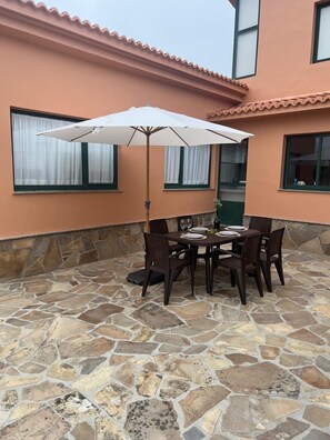 Een terras buiten met een stenen vloer, een tafel met stoelen en een witte parasol.