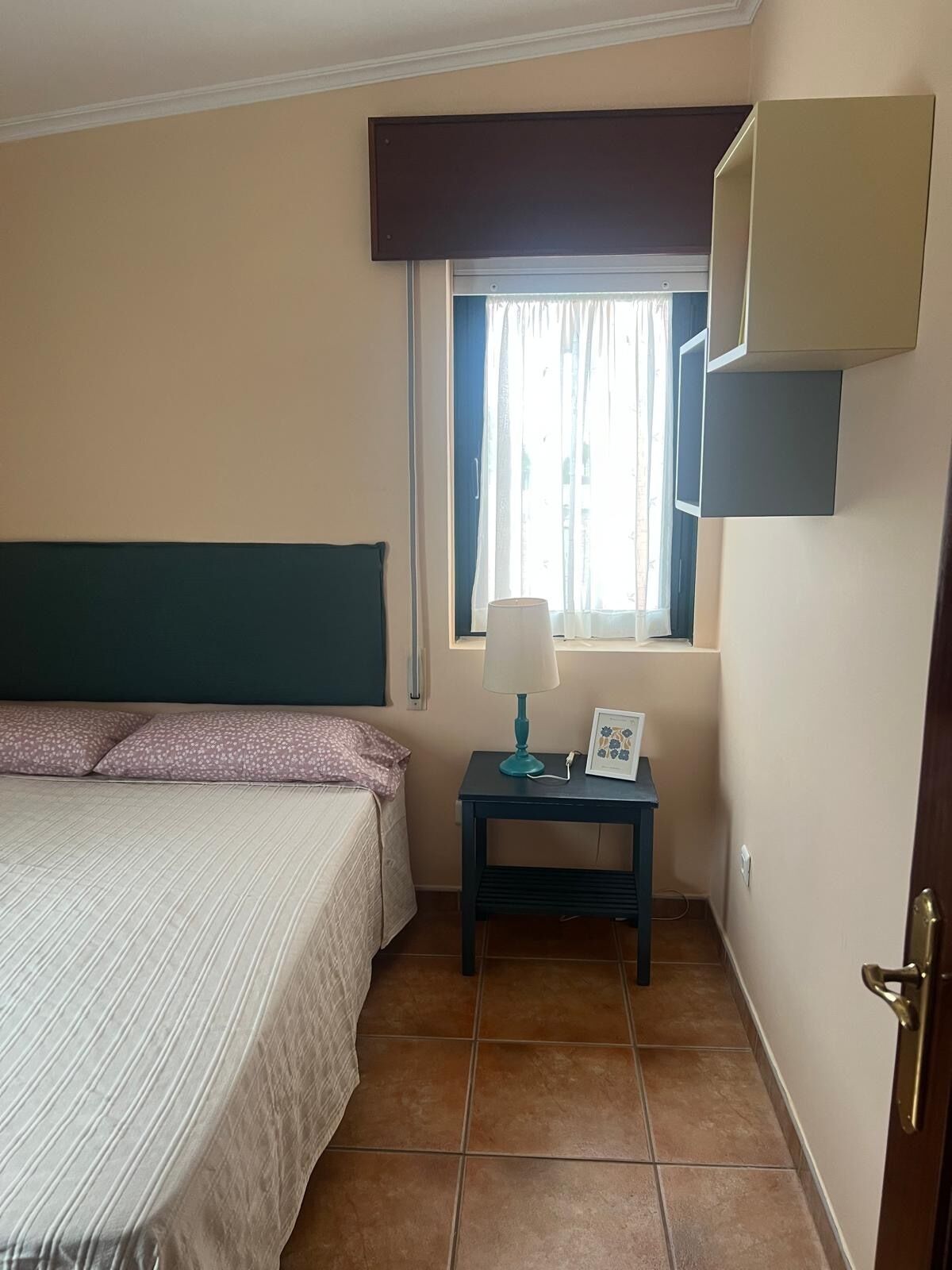 A bedroom with a bed, a nightstand with a lamp, a window with curtains, and a door.