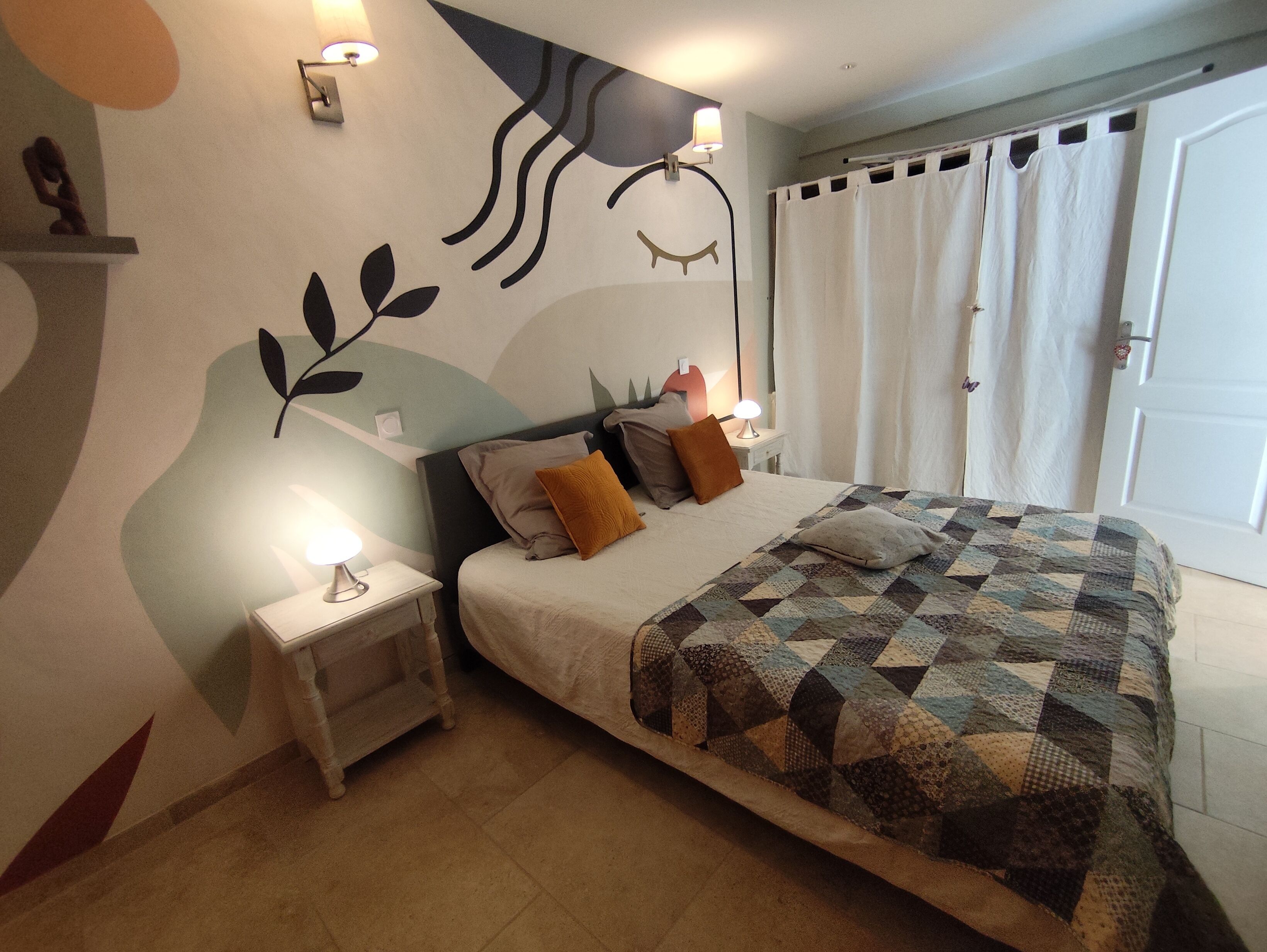 A bedroom with a bed, nightstands, a wall mural, and a door with curtains.