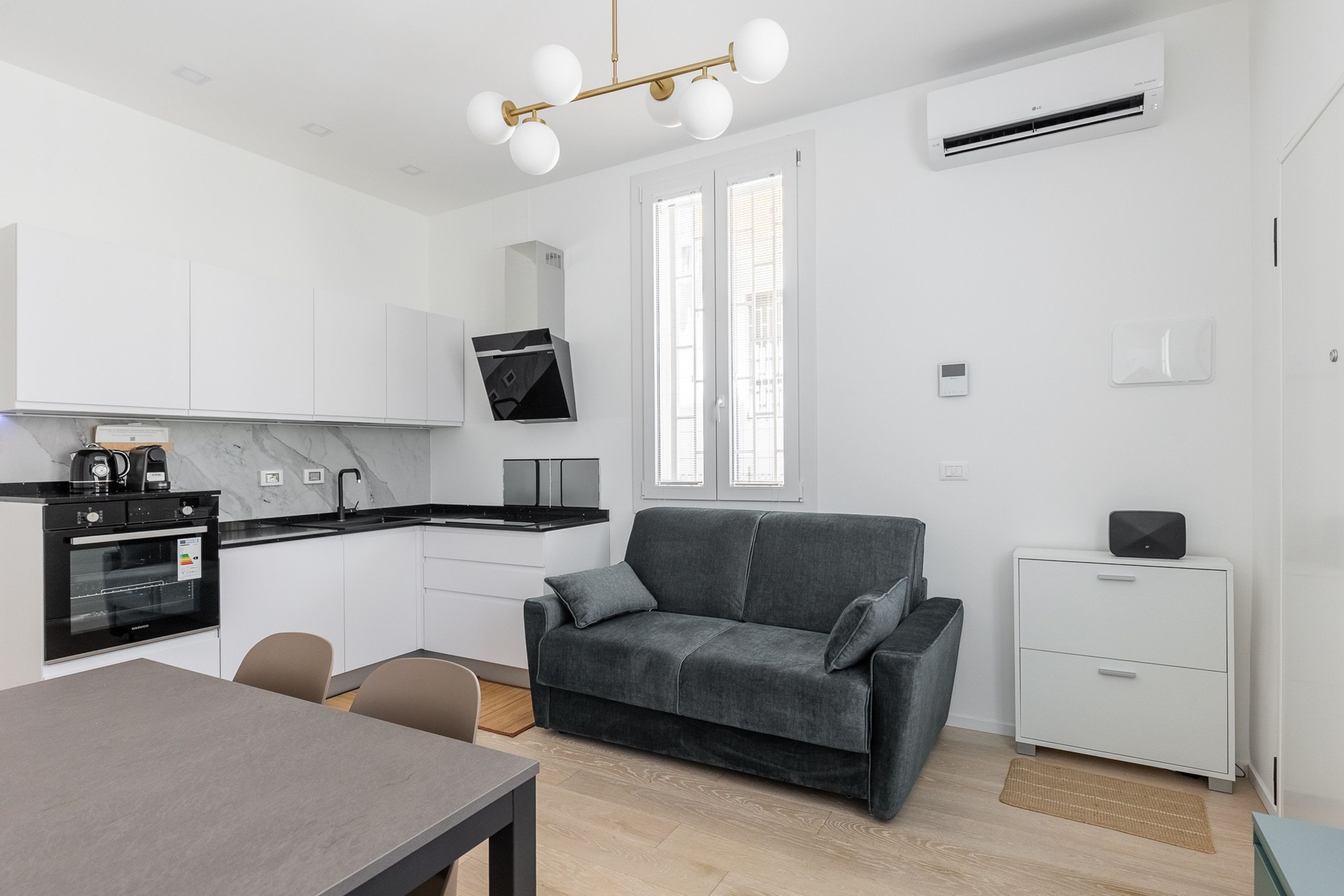 Une cuisine moderne dotée d’un four encastré, d’une table à manger avec des chaises, d’un canapé gris et d’une petite armoire blanche.