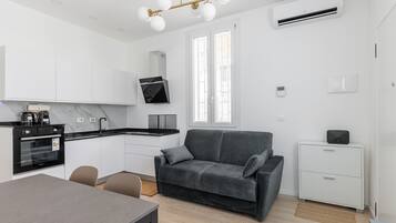 A modern kitchen with a built-in oven, a dining table with chairs, a grey sofa, and a small white cabinet.