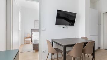 A modern dining area with a table and chairs, a flat-screen TV mounted on the wall, and a view into a bedroom through an open door.