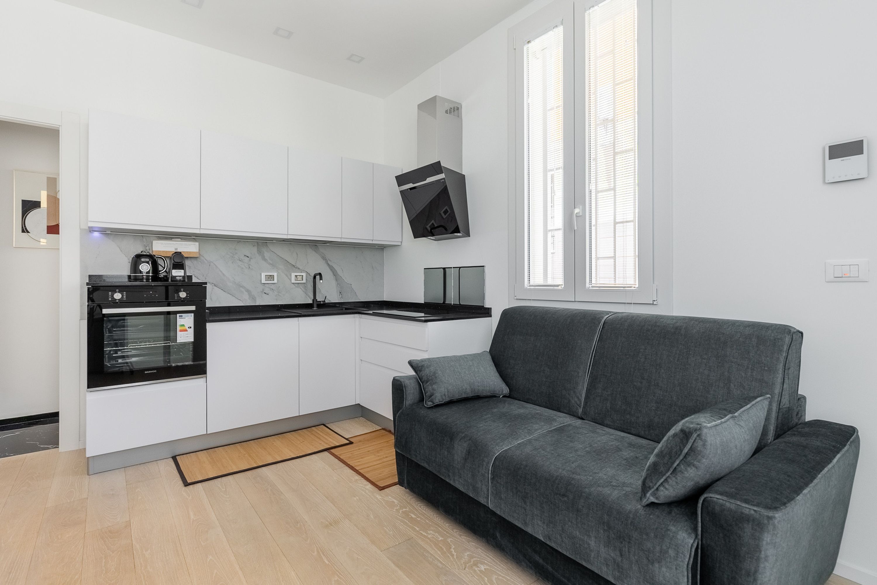 A modern kitchen with white cabinets, a black oven, and a grey sofa in a living area.