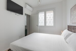 Un dormitorio con cama, televisión montada en la pared, ventana con persianas y una puerta.
