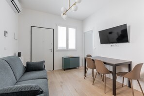 A modern living room with a grey sofa, a dining table with chairs, a flat-screen TV mounted on the wall, and a window with blinds. - La Corte Vermena by Wonderful Italy (Bologna)