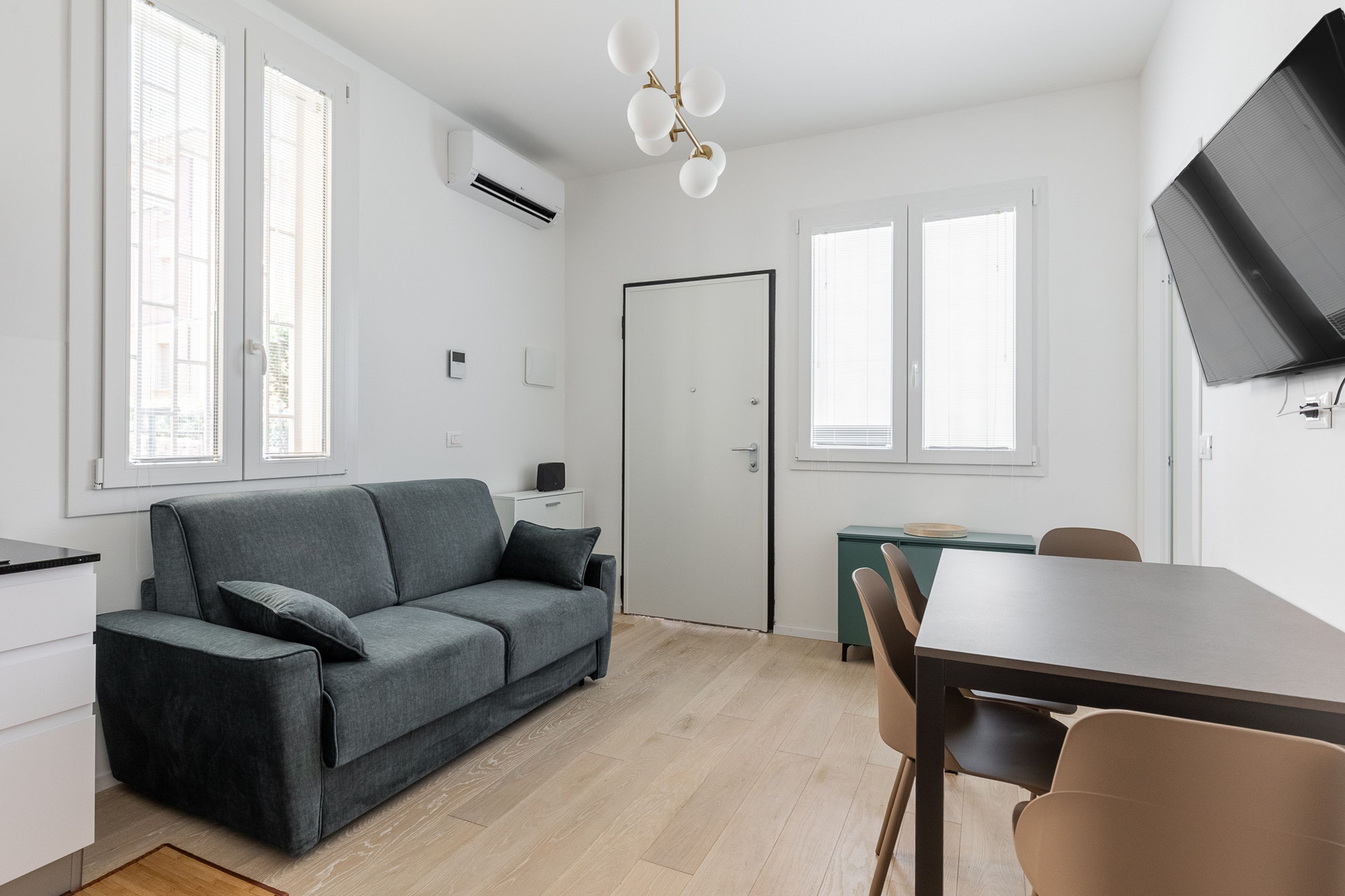 A modern, well-lit room with a grey sofa, a dining table, and a TV mounted on the wall.