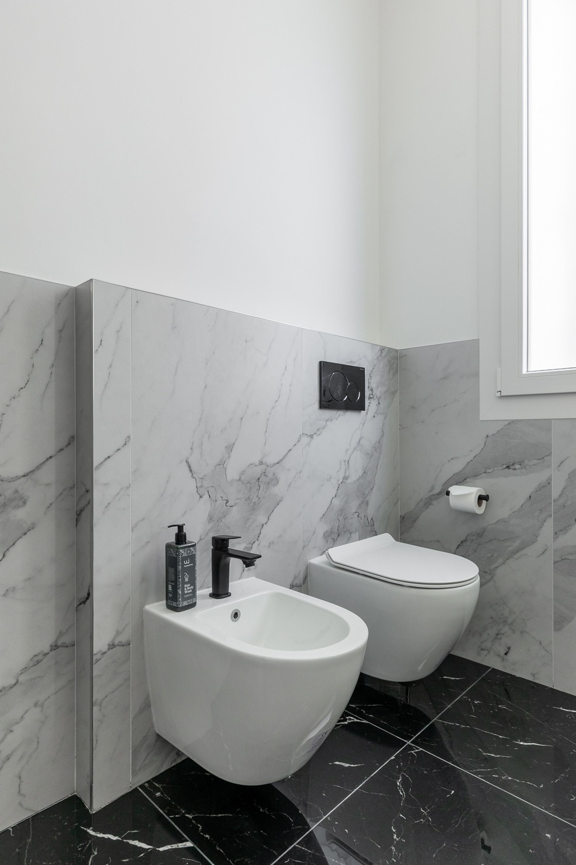 A bathroom with a bidet, a toilet, a soap dispenser, and a toilet paper holder on the wall.