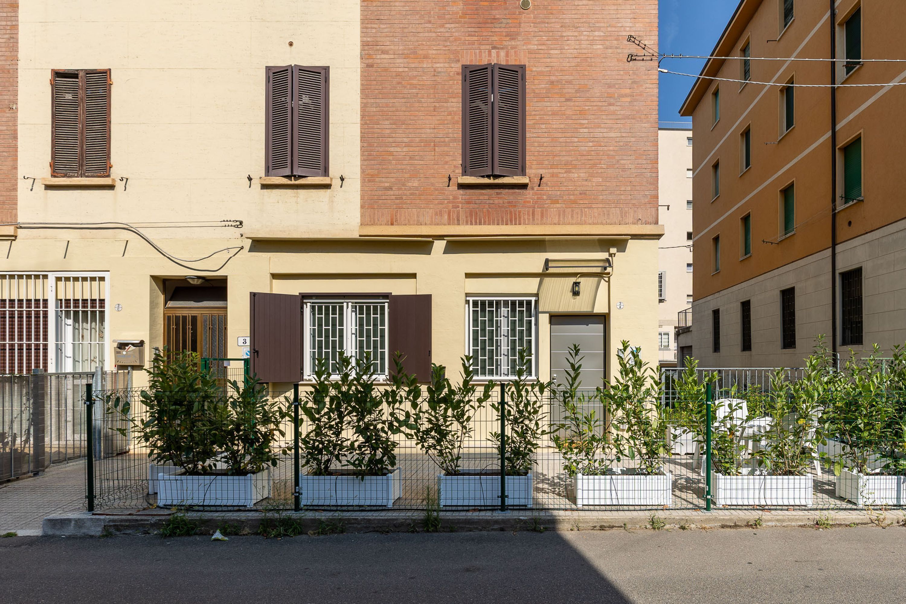 A two-story building with a beige facade, brown shutters, and a metal fence with potted plants.