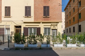 Ein zweistöckiges Gebäude mit beiger Fassade, braunen Fensterläden und einem Metallzaun mit Blumentöpfen.