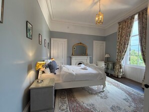 Een slaapkamer met een bed, nachtkastje, spiegel, open haard en een raam met gordijnen.