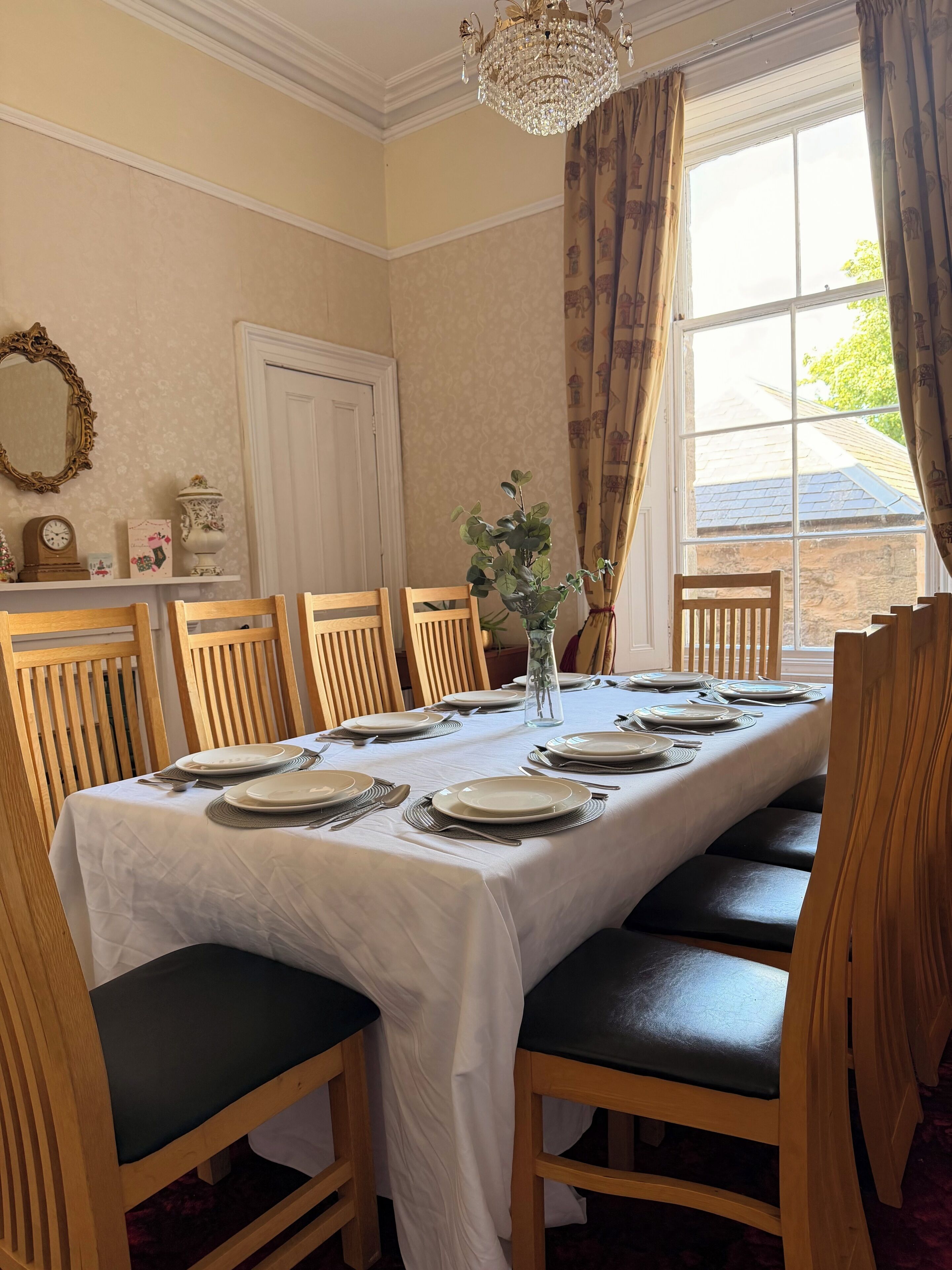 A dining room with a long table set for six, wooden chairs with green cushions, a chandelier, and a window with curtains.