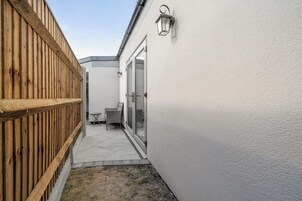 Um corredor exterior estreito, com uma cerca de madeira de um lado e uma parede branca do outro, que leva a uma entrada com porta de vidro.