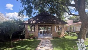 A gazebo with a brick structure, a paved pathway leading to it, and a lawn area with chairs and potted plants.