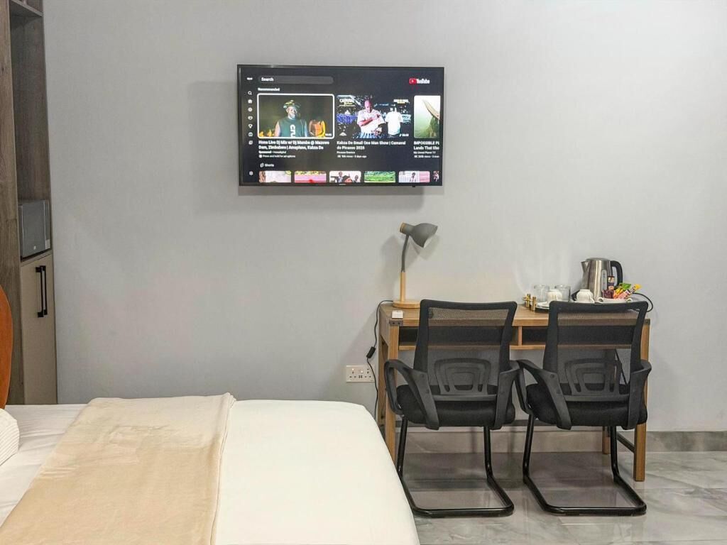 A hotel room with a bed, a desk with two chairs, a TV mounted on the wall, and a lamp.