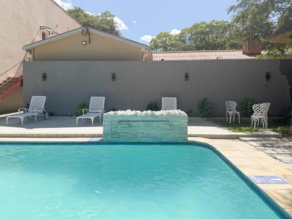 A swimming pool with a stone accent wall, white chairs, and a covered area with stairs.