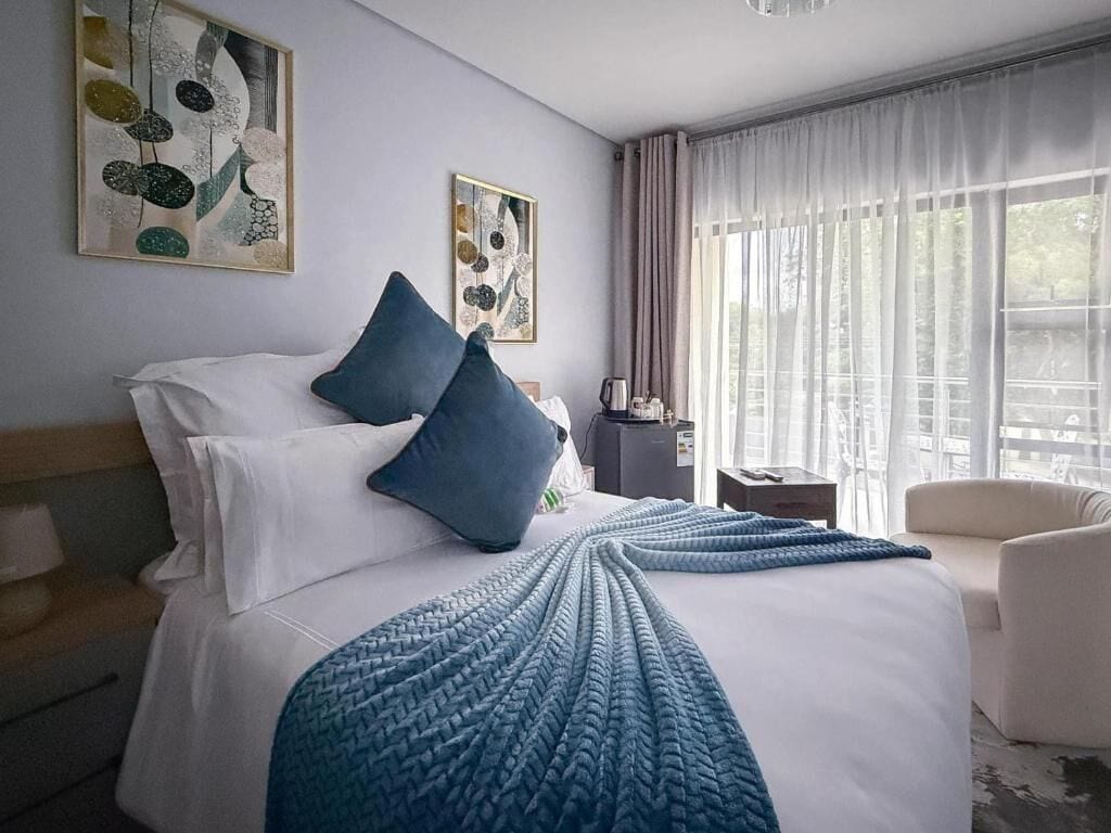Habitación de hotel con cama, almohadas, colcha, cafetera, sillón y cuadros en las paredes.