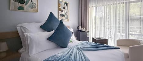 Habitación de hotel con cama, almohadas, colcha, cafetera, sillón y cuadros en las paredes.