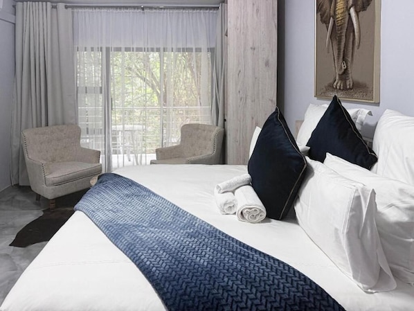 Quarto de hotel com uma cama grande, duas poltronas, uma janela com cortinas e uma obra de arte pendurada na parede.