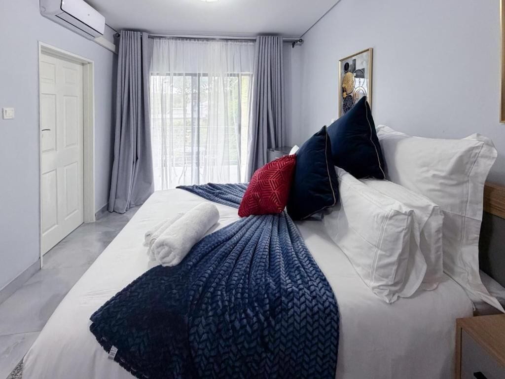 Hotellihuone, jossa on sänky, ikkuna verhoilla, ovi ja seinälle asennettu ilmastointilaite.