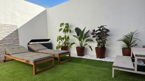 Et moderne udendørsareal med kunstgræs, træmøbler og krukker med planter foran en hvid væg.