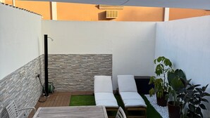 En terrasse med et træbord og stole, et lille siddeområde med puder og krukker med planter.