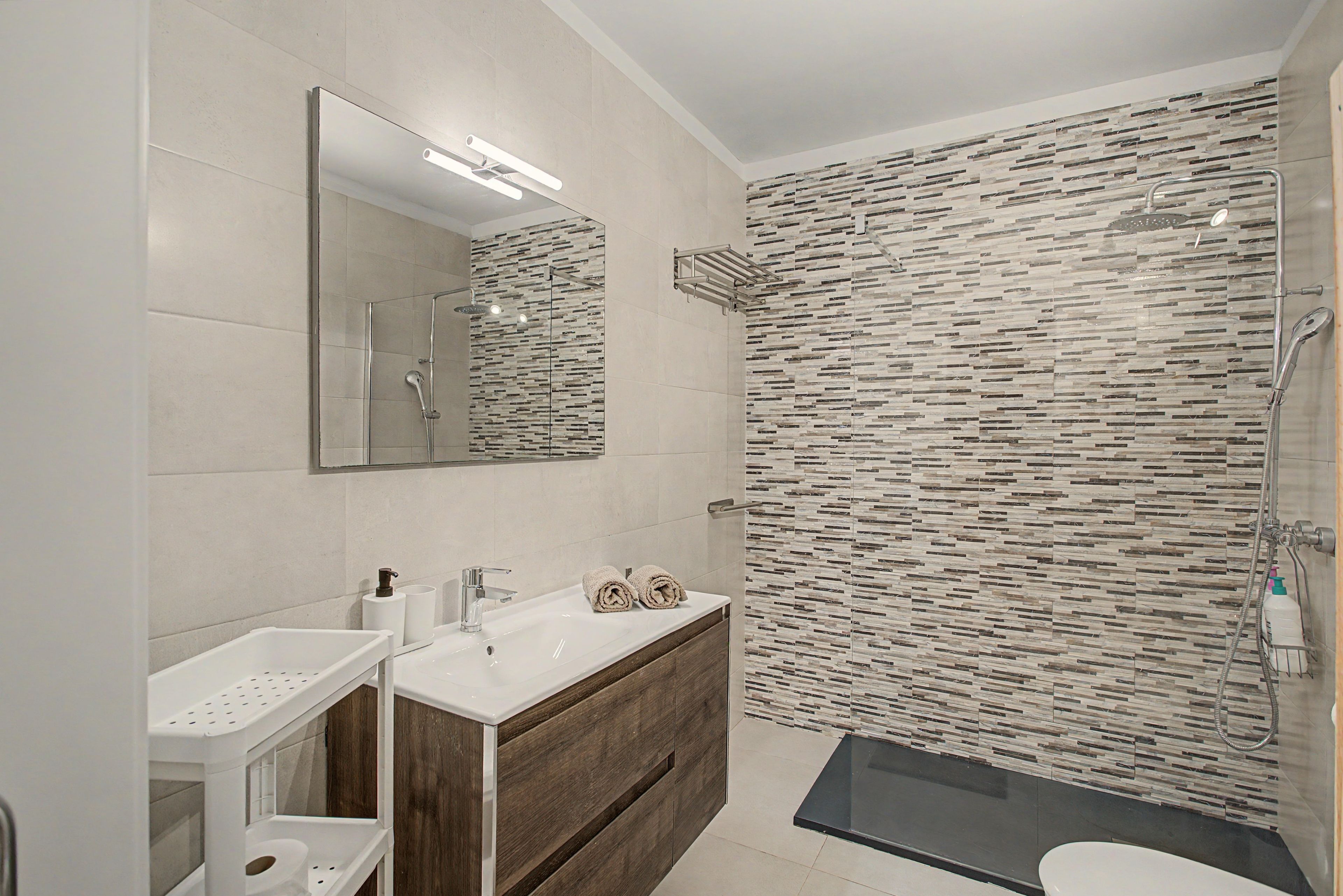 Een moderne badkamer met een douche, een wastafel met twee kommen, een spiegel en een houten opbergmeubel.