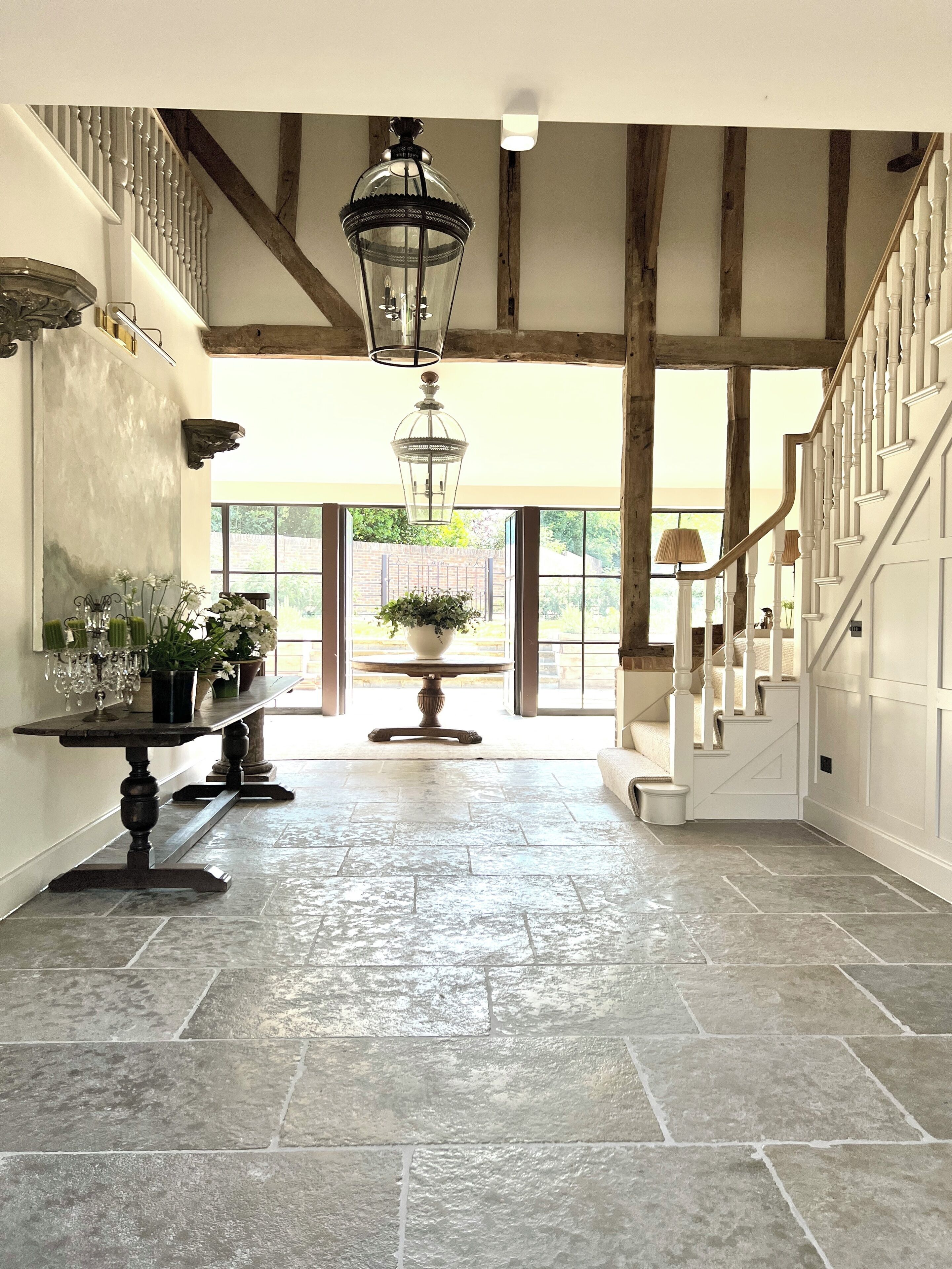 A spacious hallway with a stone floor, a grand staircase with white balustrades, and a large window offering a view of greenery outside.