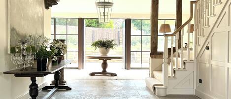 A spacious hallway with a stone floor, a grand staircase with white balustrades, and a large window offering a view of greenery outside.