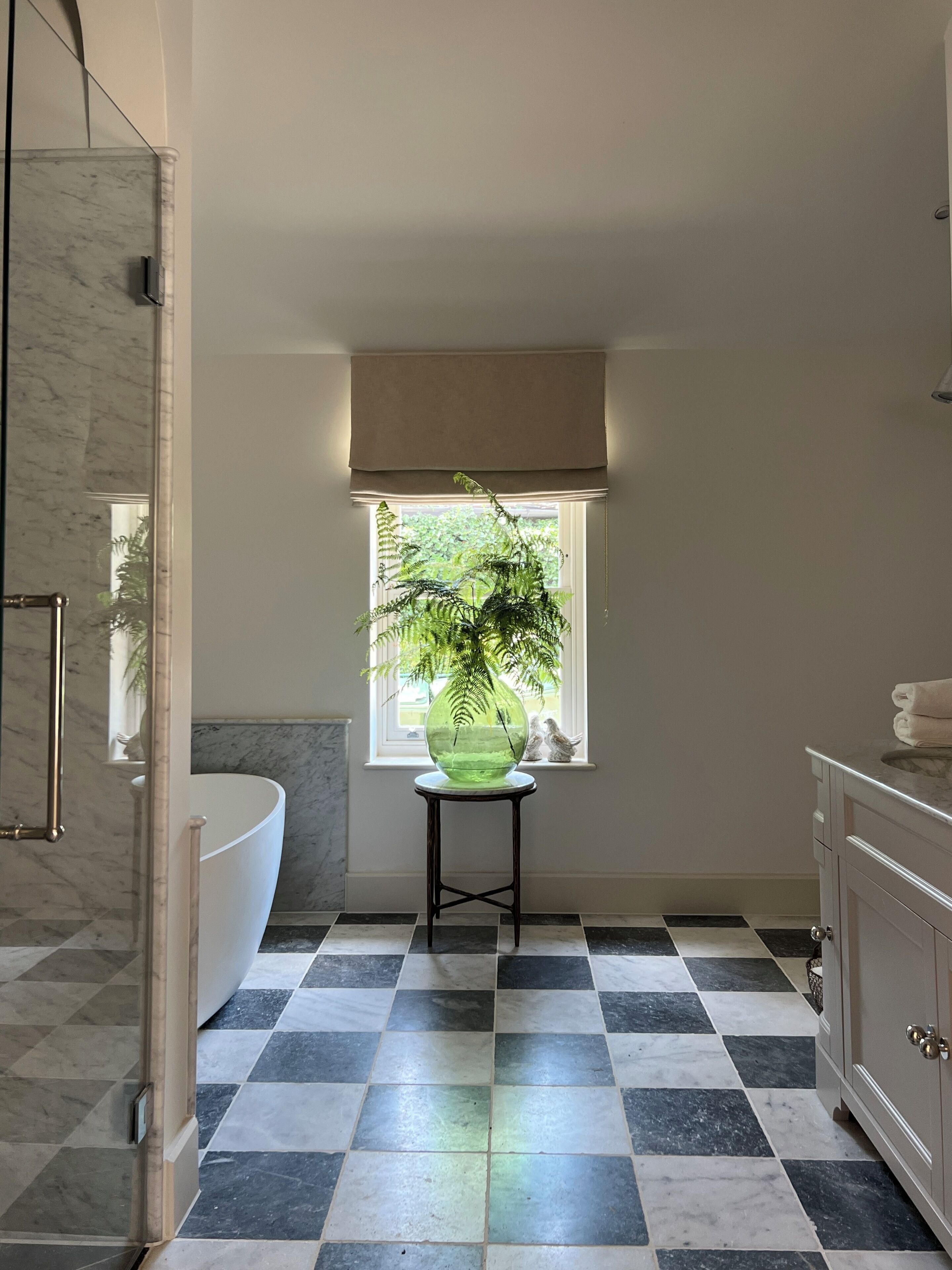 A bathroom with a checkered floor, a large window with a view of greenery, a glass vase with a plant, and a bathtub.