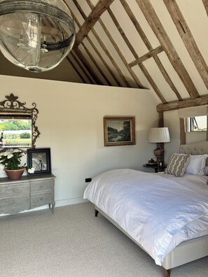 A bedroom with a large bed, a wooden ceiling, a chandelier, a dresser, a mirror, and a window.