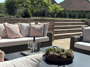 Un espace extérieur aménagé avec des meubles en osier, des coussins et un plateau contenant des verres et une bouteille.