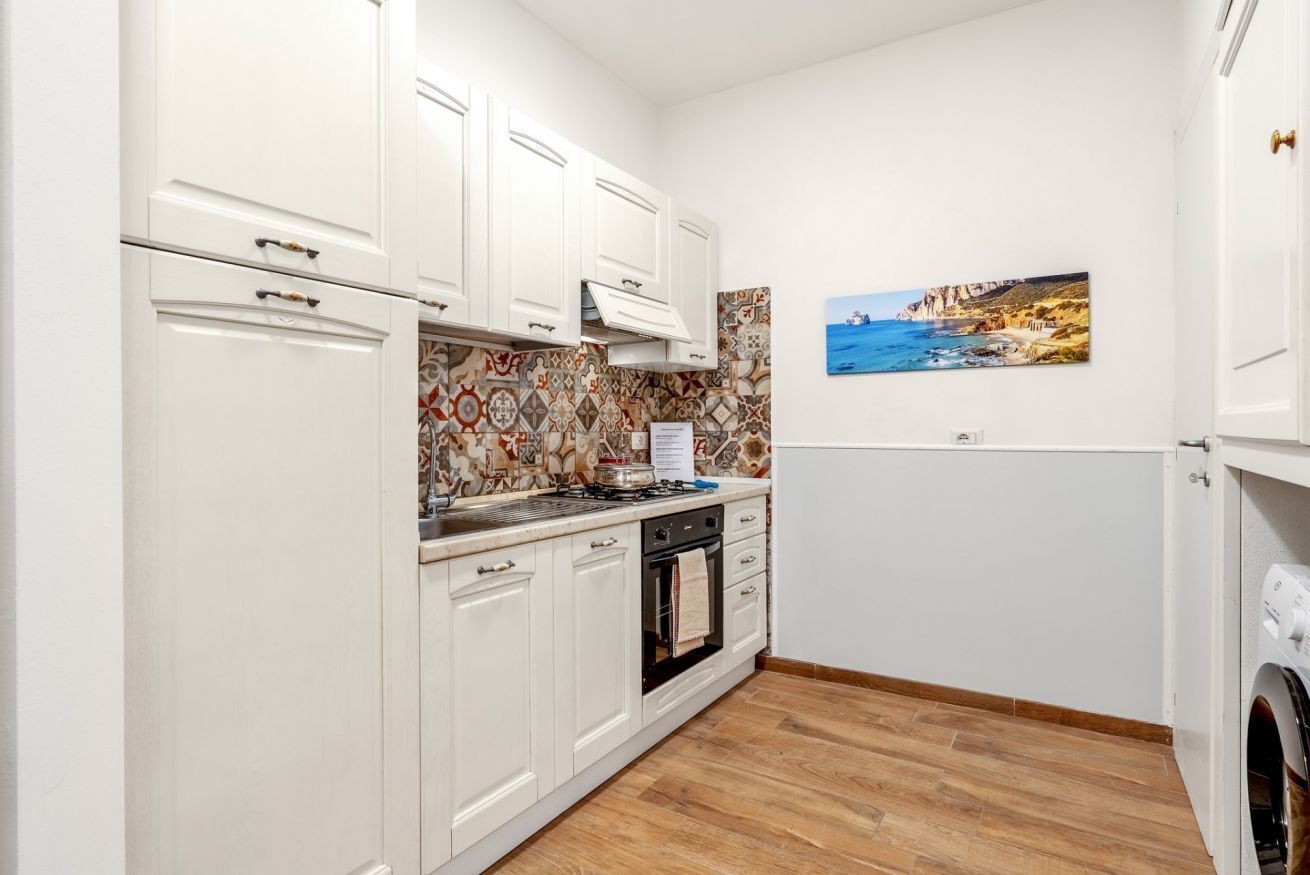 A kitchen with white cabinets, a stove, and an oven. There is a patterned backsplash and a painting of a coastal scene on the wall.