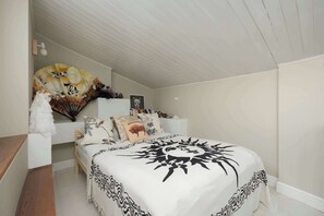 Ein Schlafzimmer mit einem Bett, einem Nachttisch, einer Lampe, einem Ventilator und einer Wand mit einem Gemälde.