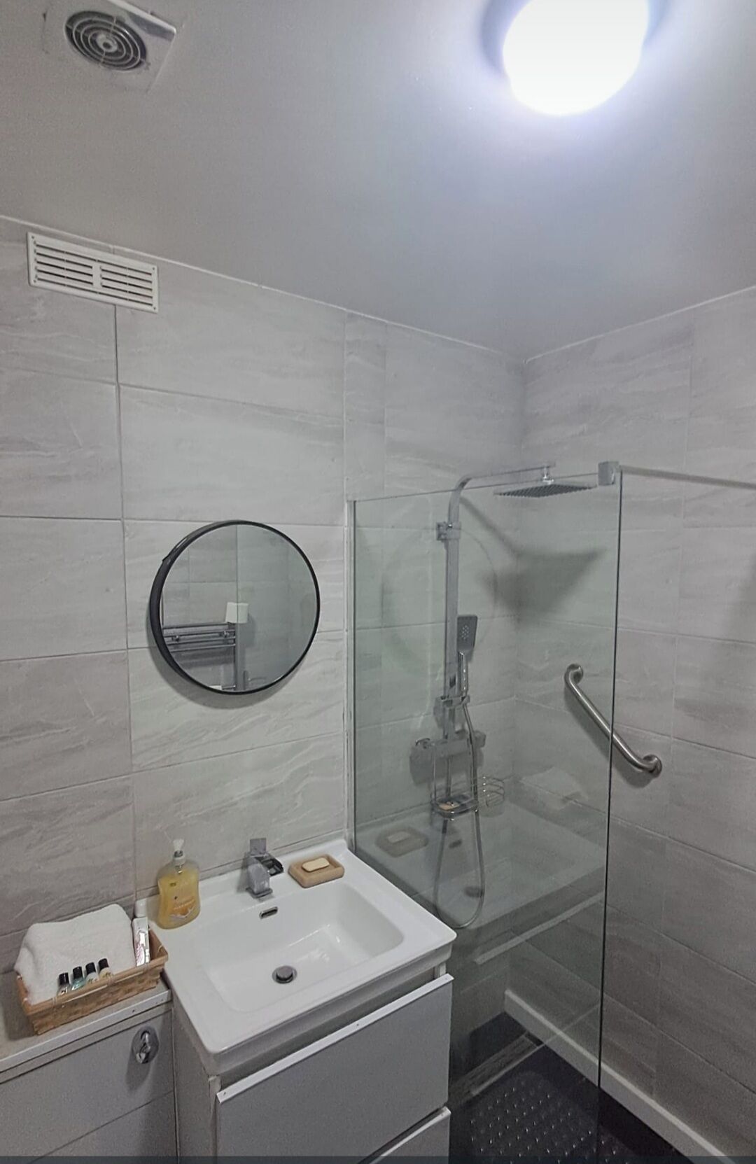 Un baño moderno con cabina de ducha de vidrio, un lavamanos blanco con espejo y un pequeño mueble de almacenamiento de madera.