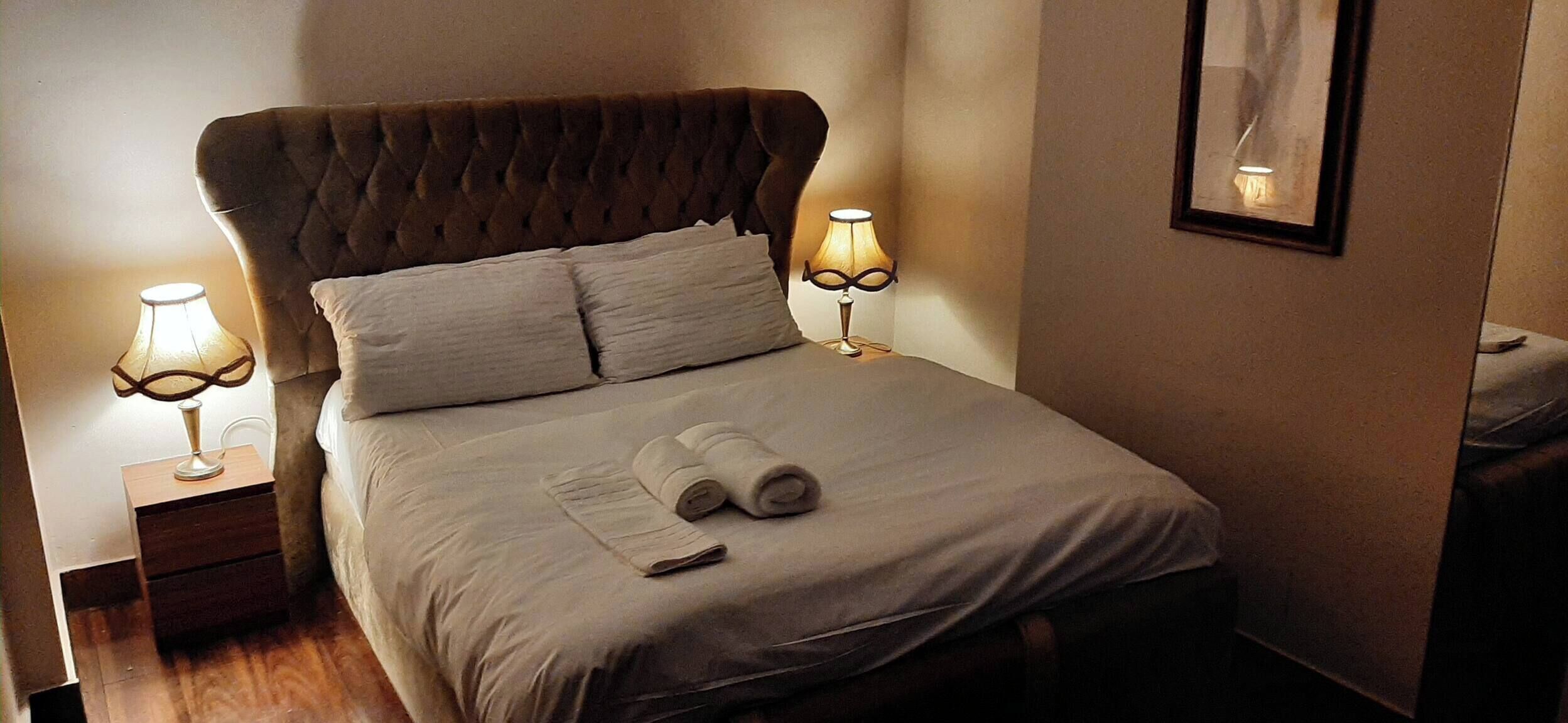 Una cama bien hecha con cabecera, dos lámparas de noche y una toalla enrollada sobre la cama.