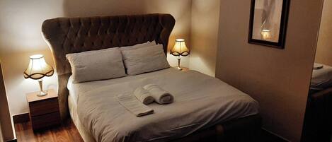 Una cama bien hecha con cabecera, dos lámparas de noche y una toalla enrollada sobre la cama.