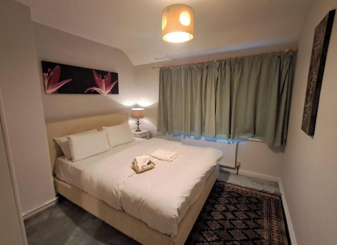 Un dormitorio con cama, mesita de noche, lámpara, ventana con cortinas y un cuadro en la pared.