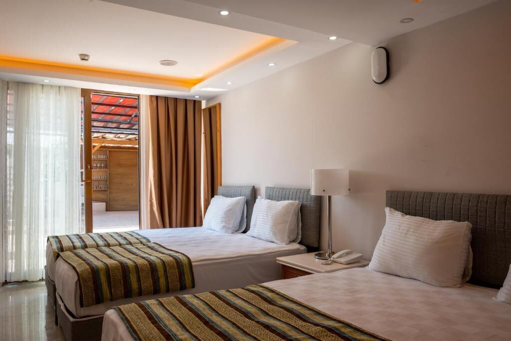 Quarto de hotel com duas camas, uma mesa de cabeceira com abajur, telefone e vista para uma varanda com cortinas.