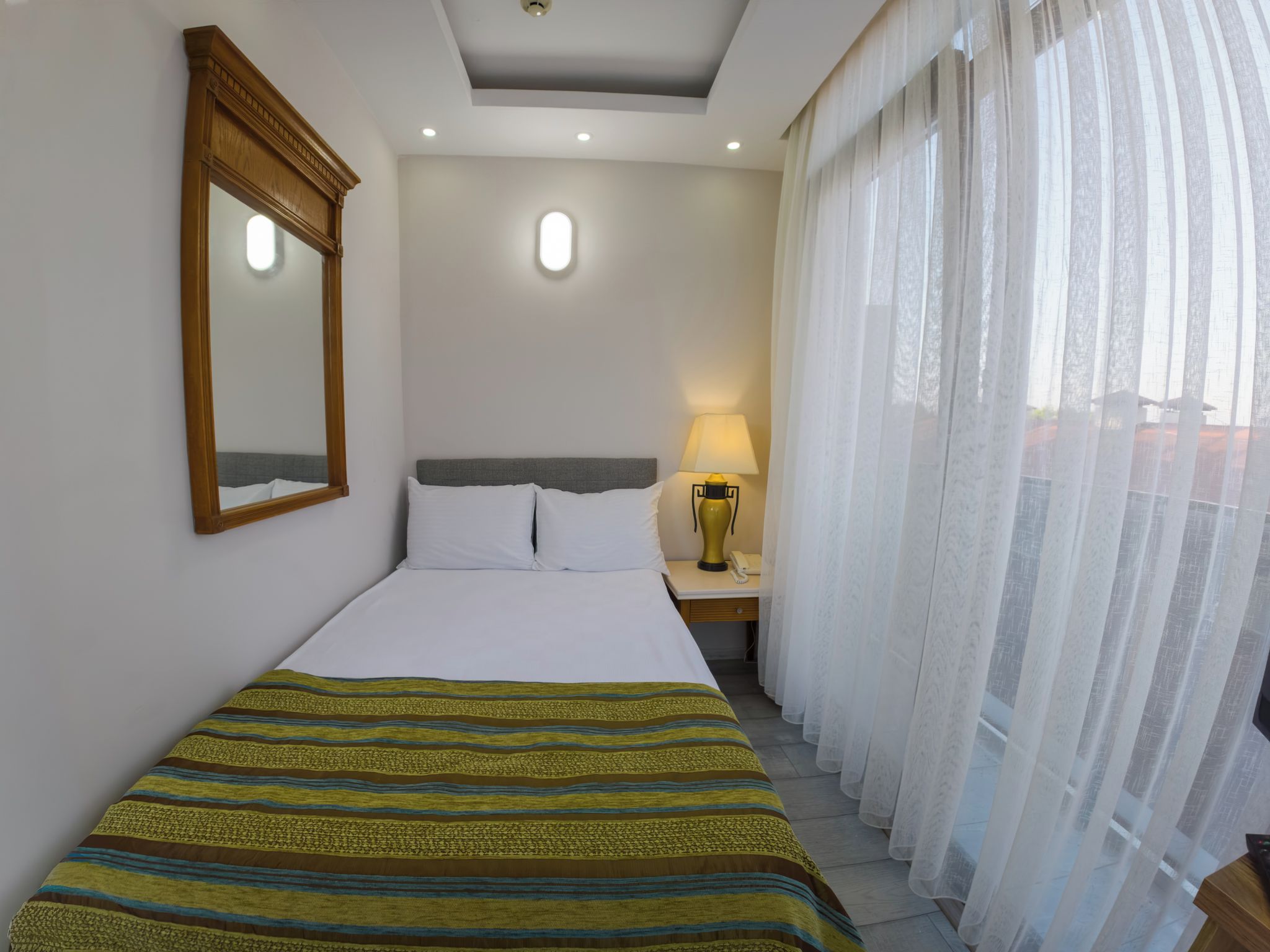 Een hotelkamer met een bed, nachtkastje, lamp, spiegel en dunne gordijnen.