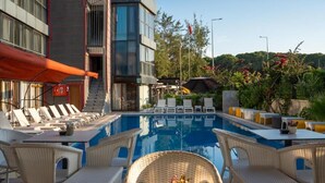 A swimming pool with lounge chairs and tables, surrounded by a modern building with a glass facade. - Velaris Boutique Hotel (Serik)