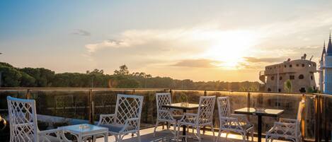An outdoor seating area with tables and chairs, overlooking a body of water and a distant building with a tower.
