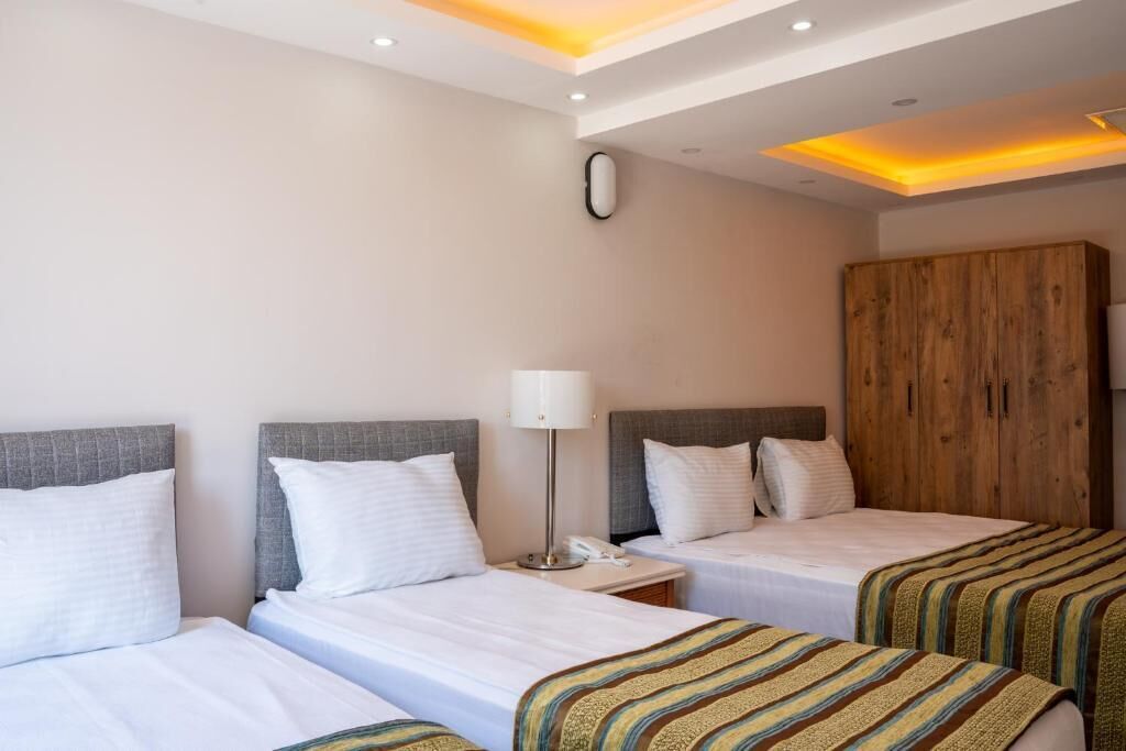 A hotel room with two single beds, a wooden wardrobe, a bedside table with a lamp, and a ceiling light fixture.